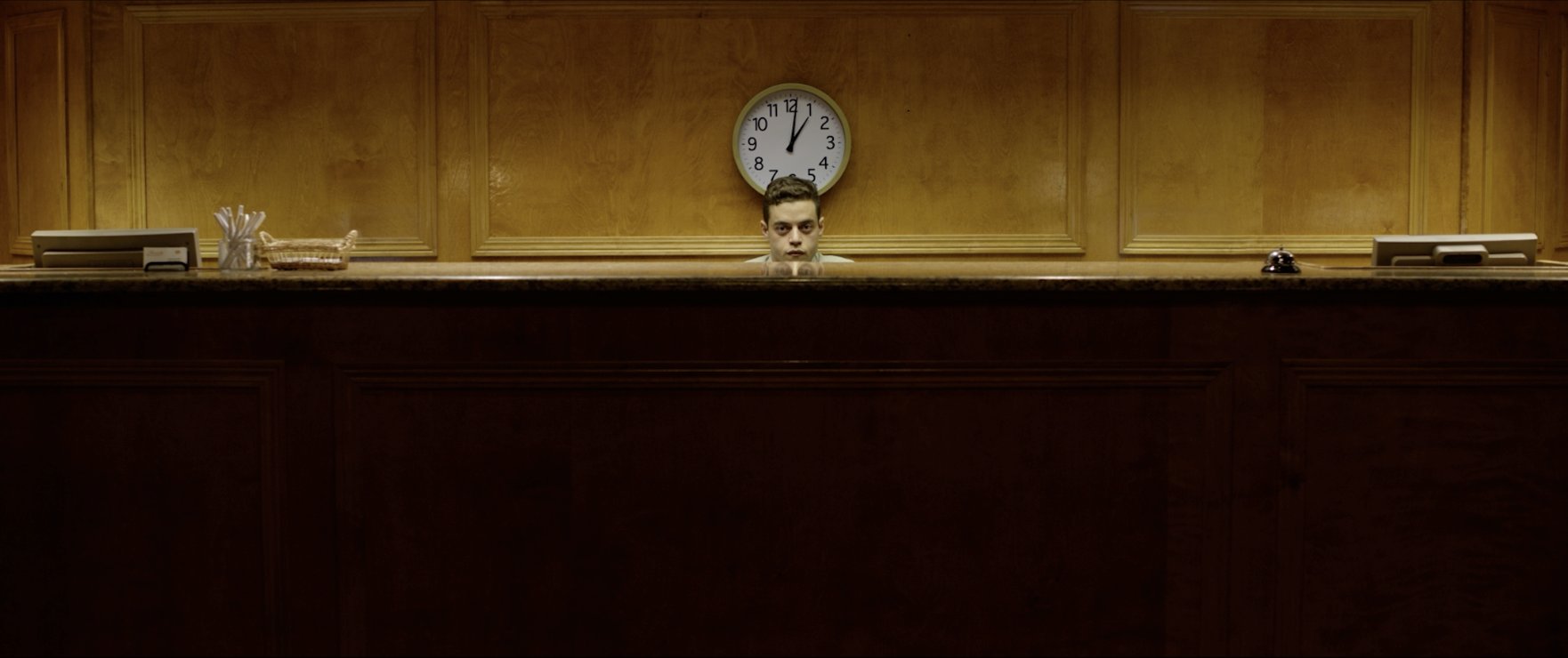 Still from the reel: a man peers over a wood-paneled hotel reception desk beneath a wall clock